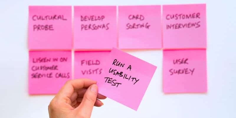 hand-holding-pink-sticky-notes-with-text-run-a-usability-test-with-seven-more-sticky-notes-on-the-white-board-all-related-to ux-design-process